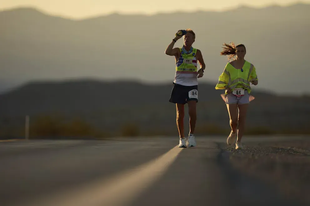 Danny Westergaard corre al atardecer por una carretera del desierto junto a una corredora de su equipo, ambos con chaleco reflectante y el dorsal 84