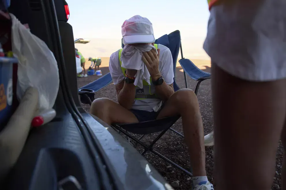 Un corredor agotado descansa en una silla plegable junto al vehículo de apoyo, cubriéndose la cara con un paño húmedo para combatir el calor